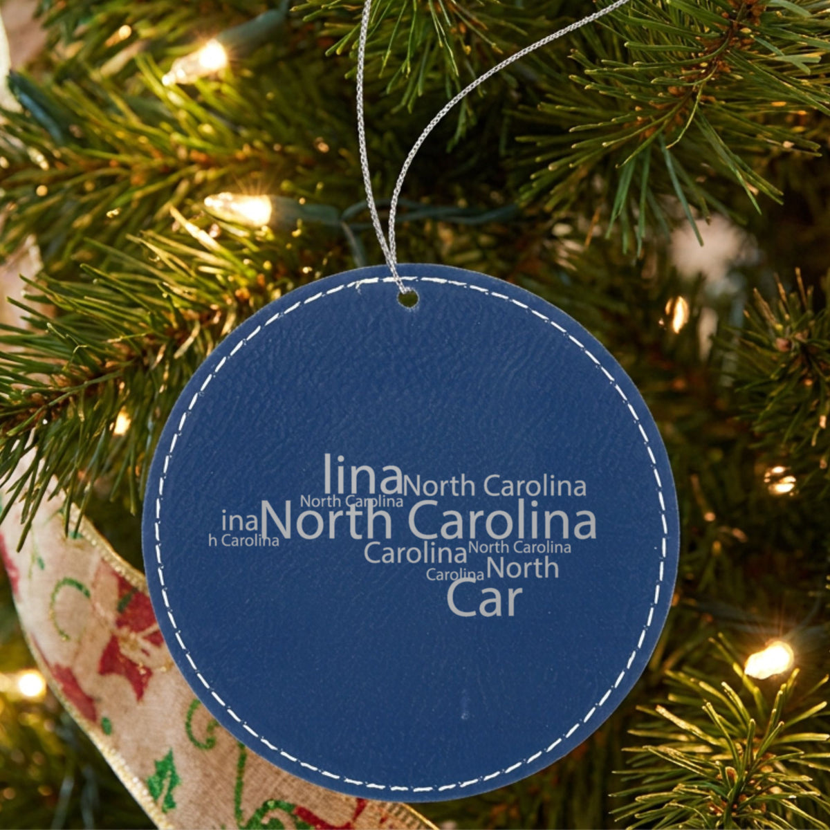Blue leatherette round Christmas ornament laser engraved with the North Carolina Word Cloud design, hanging on a tree