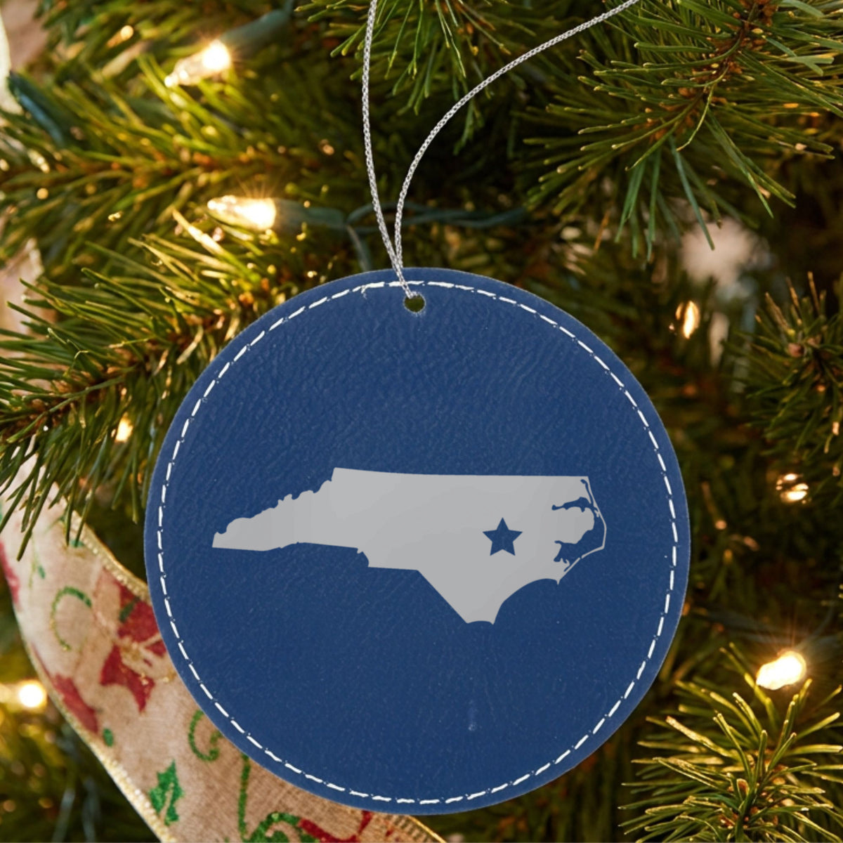 Blue leatherette round Christmas ornament laser engraved with the North Carolina Star Accent design, hanging on a tree