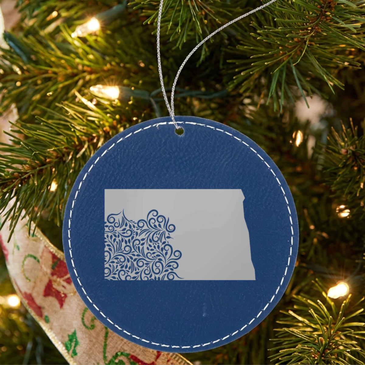 Blue leatherette round Christmas ornament laser engraved with the North Dakota Floral Damask design, hanging on a tree