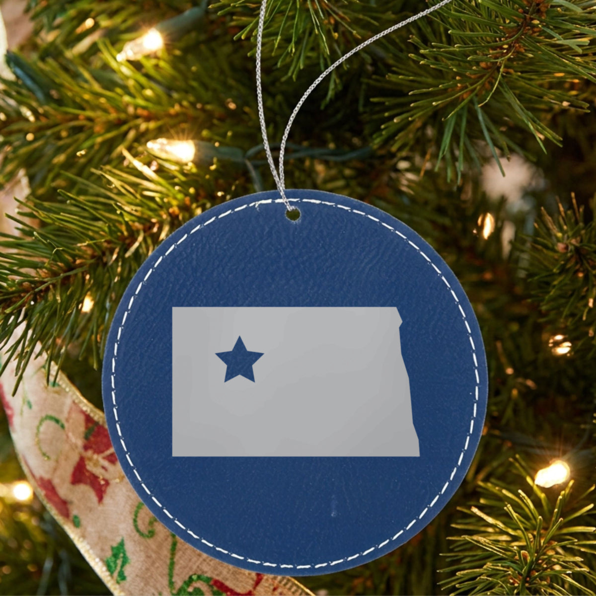 Blue leatherette round Christmas ornament laser engraved with the North Dakota Star Accent design, hanging on a tree