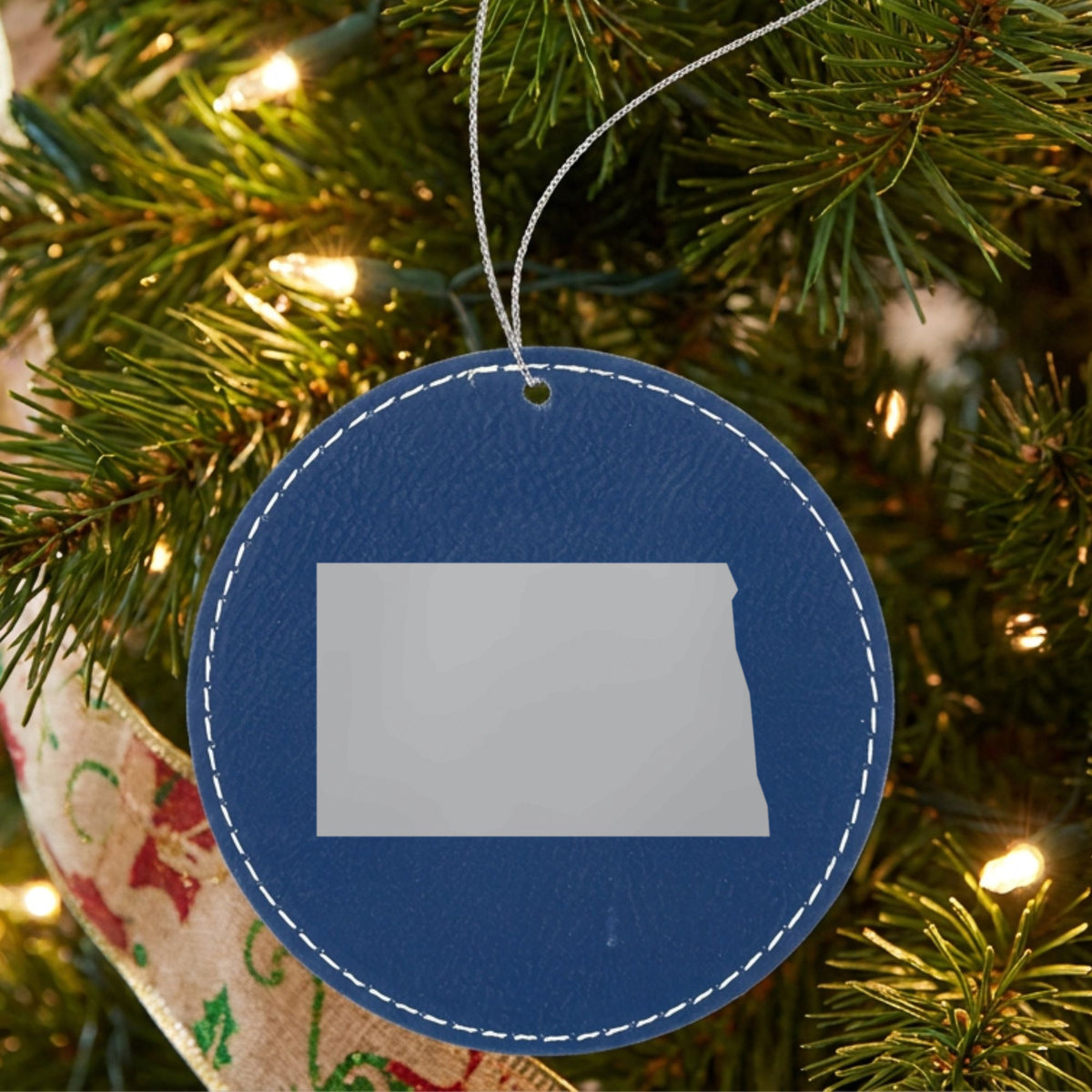 Blue leatherette round Christmas ornament laser engraved with the North Dakota Engraved State design, hanging on a tree