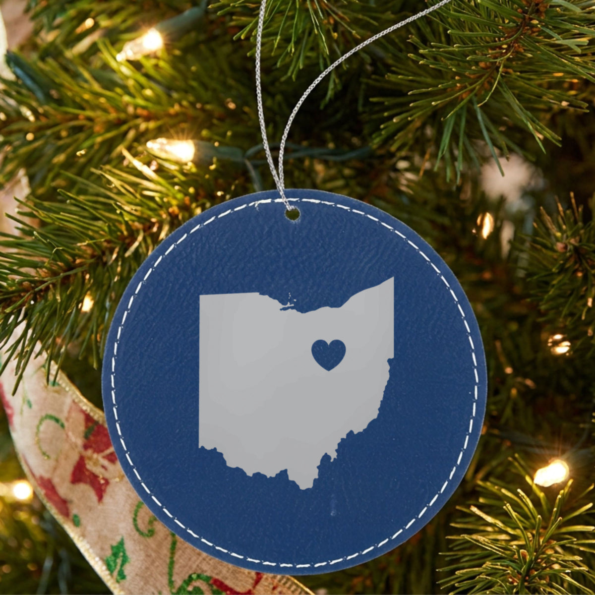 Blue leatherette round Christmas ornament laser engraved with the Ohio Heart Accent design, hanging on a tree