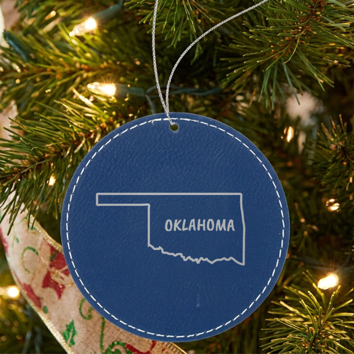 Blue leatherette round Christmas ornament laser engraved with the Oklahoma Brush Lettering design, hanging on a tree
