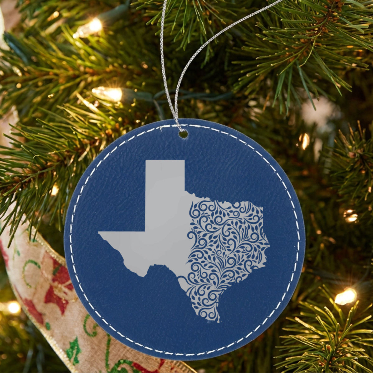 Blue leatherette round Christmas ornament laser engraved with the Texas Floral Damask design, hanging on a tree
