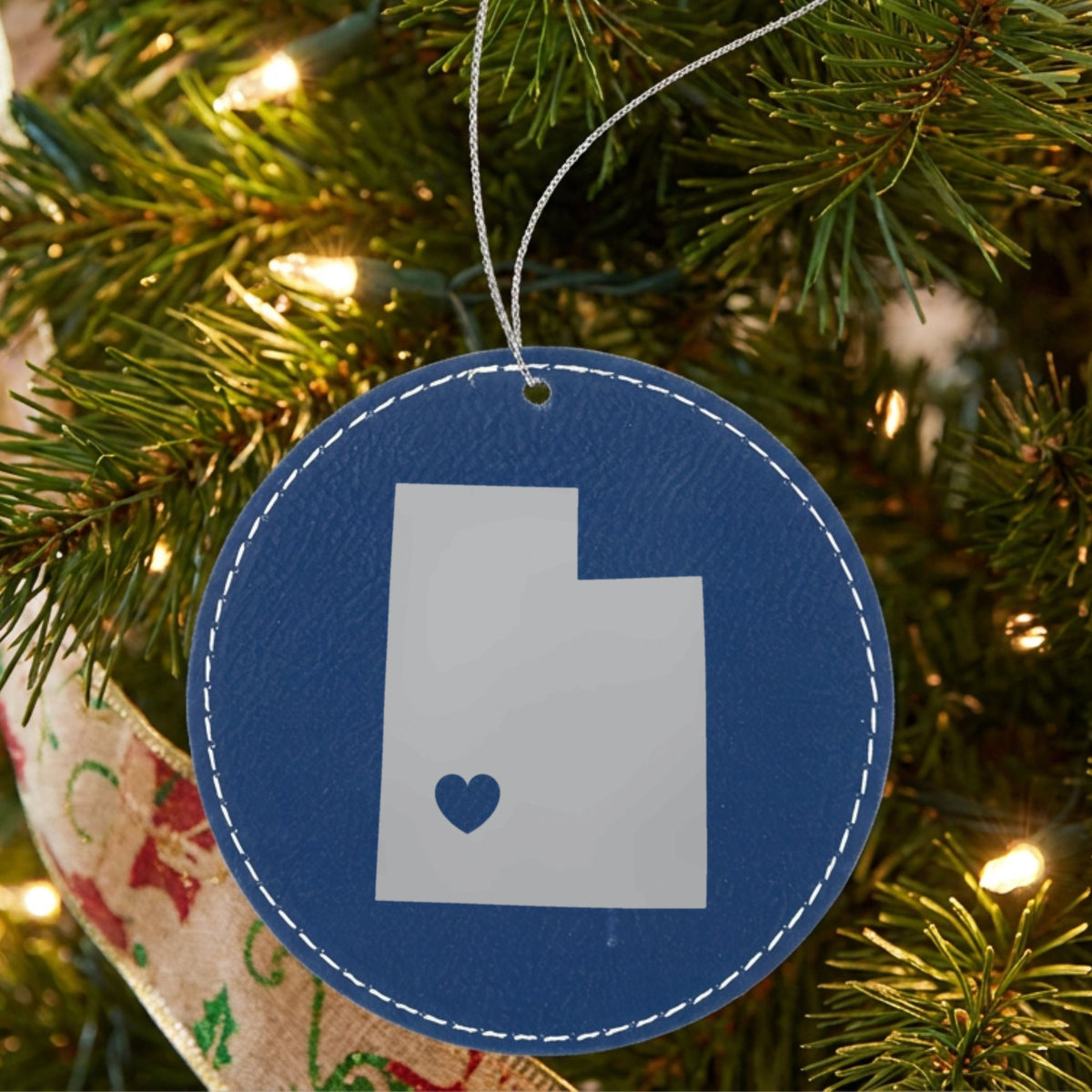 Blue leatherette round Christmas ornament laser engraved with the Utah Heart Accent design, hanging on a tree