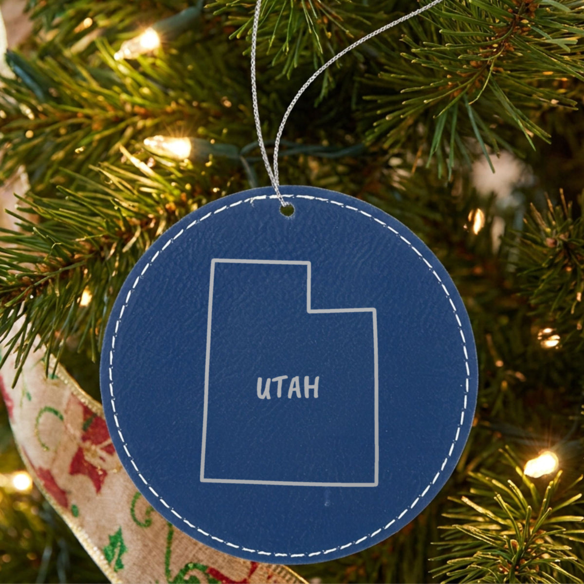 Blue leatherette round Christmas ornament laser engraved with the Utah Brush Lettering design, hanging on a tree