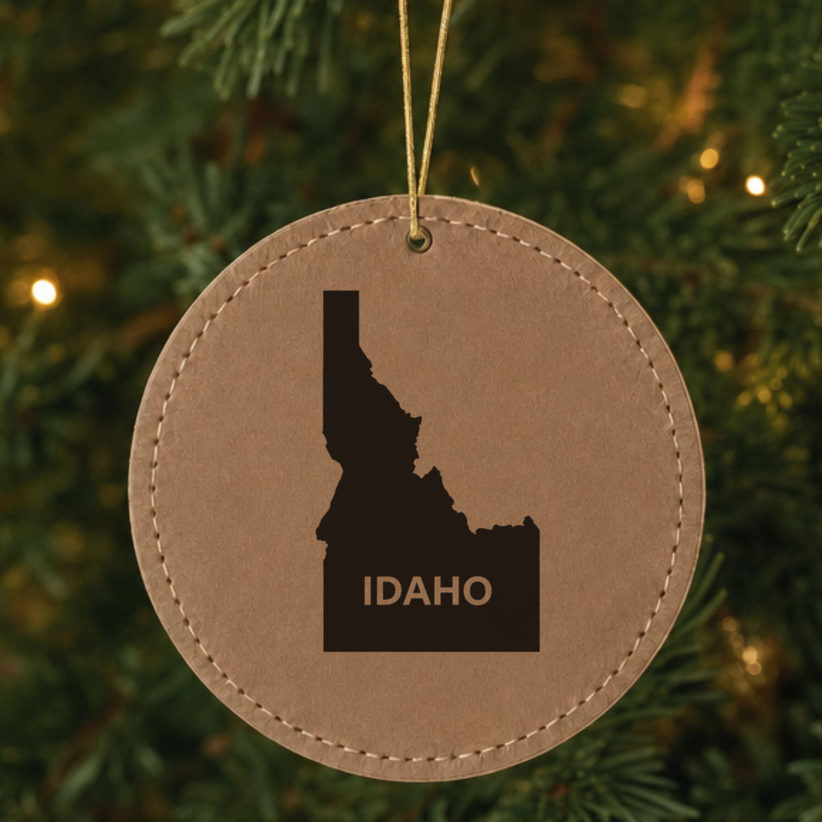 Brown leatherette round Christmas ornament laser engraved with the Idaho Bold State Name design, hanging on a tree