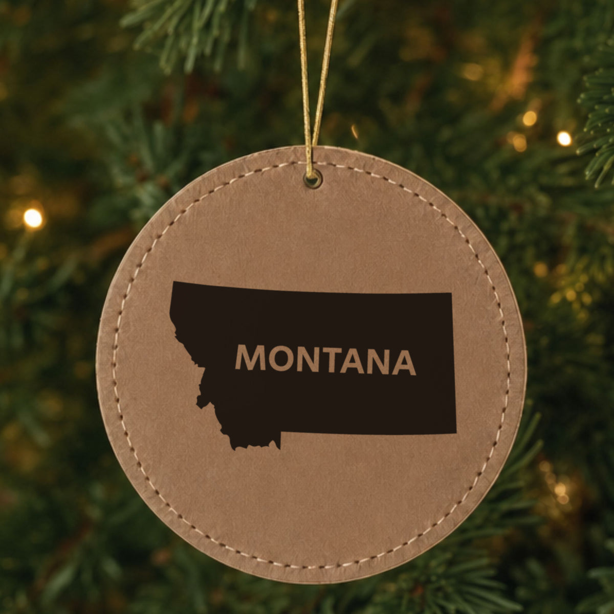 Brown leatherette round Christmas ornament laser engraved with the Montana Bold State Name design, hanging on a tree