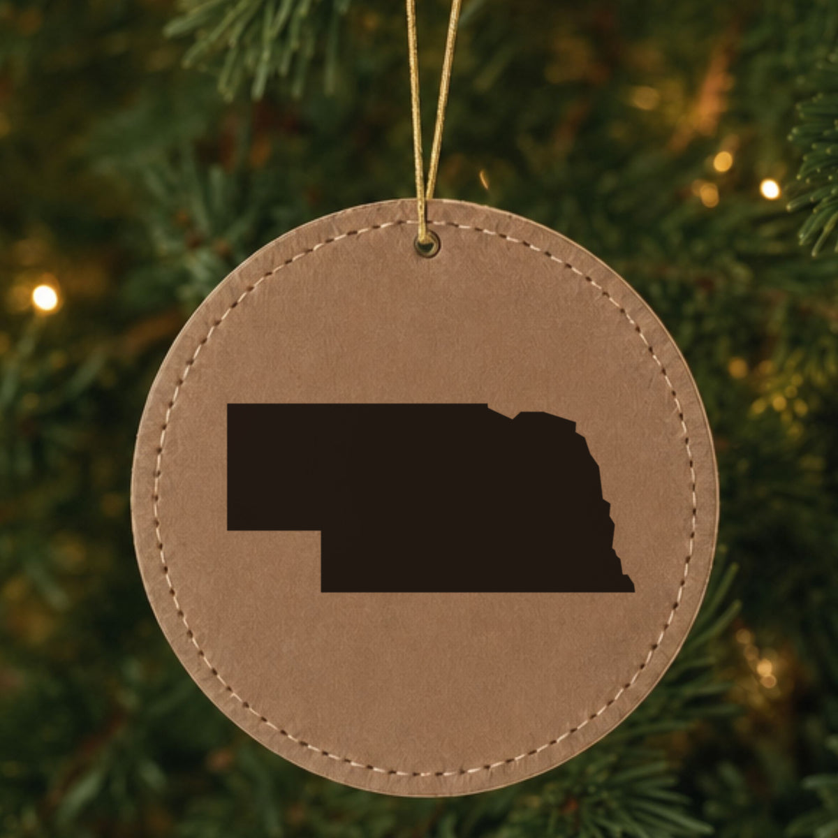 Brown leatherette round Christmas ornament laser engraved with the Nebraska Engraved State design, hanging on a tree