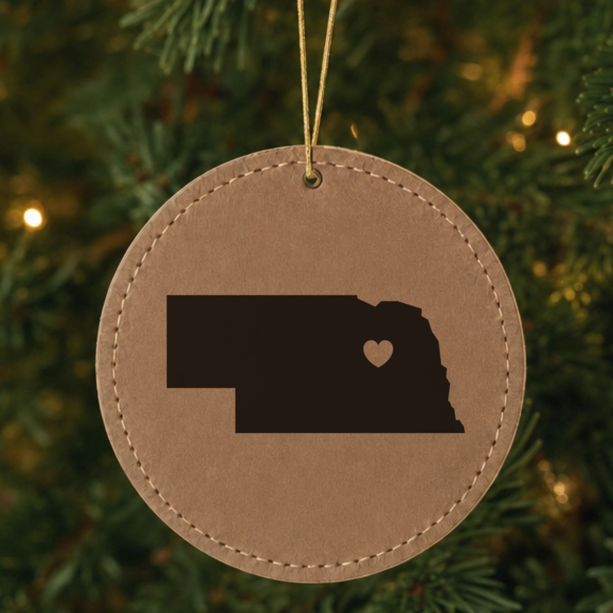 Brown leatherette round Christmas ornament laser engraved with the Nebraska Heart Accent design, hanging on a tree
