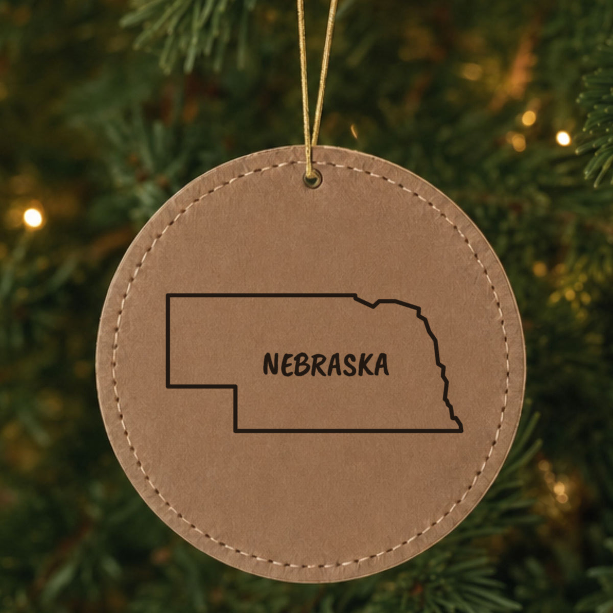 Brown leatherette round Christmas ornament laser engraved with the Nebraska Brush Lettering design, hanging on a tree