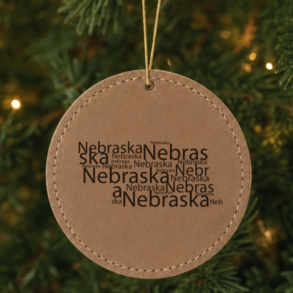 Brown leatherette round Christmas ornament laser engraved with the Nebraska Word Cloud design, hanging on a tree