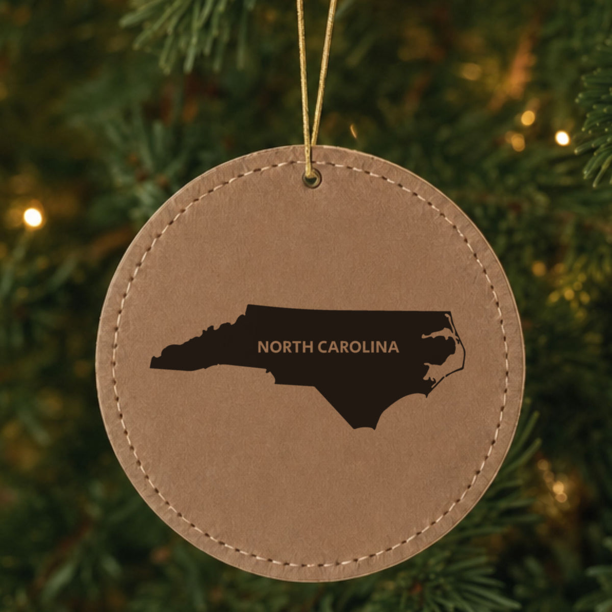 Brown leatherette round Christmas ornament laser engraved with the North Carolina Bold State Name design, hanging on a tree