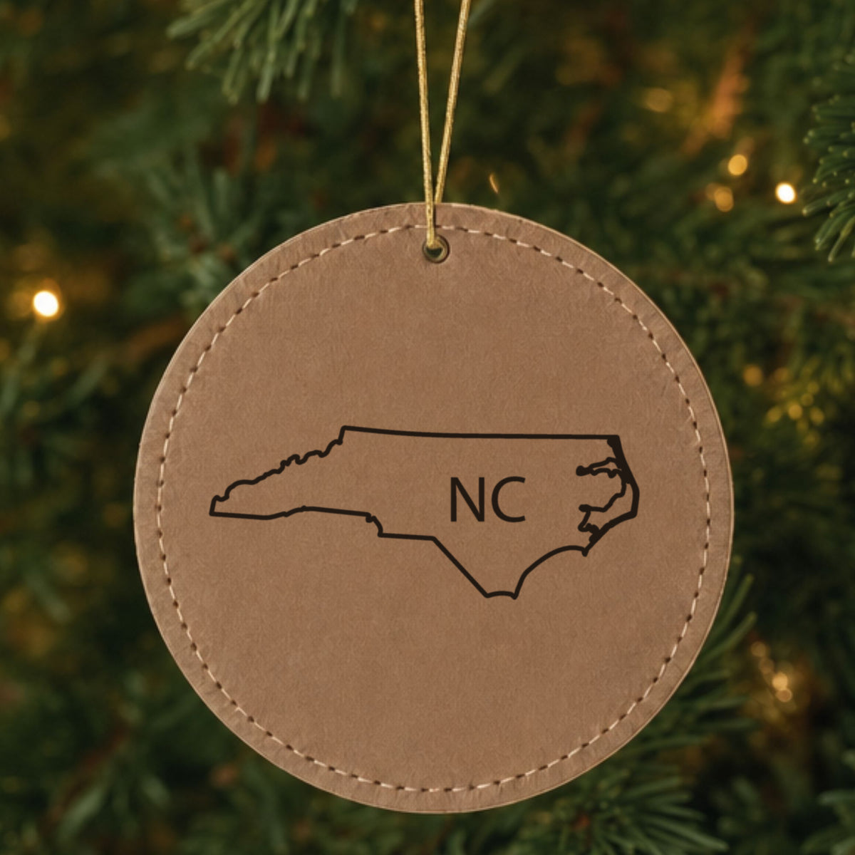Brown leatherette round Christmas ornament laser engraved with the North Carolina NC Initials design, hanging on a tree