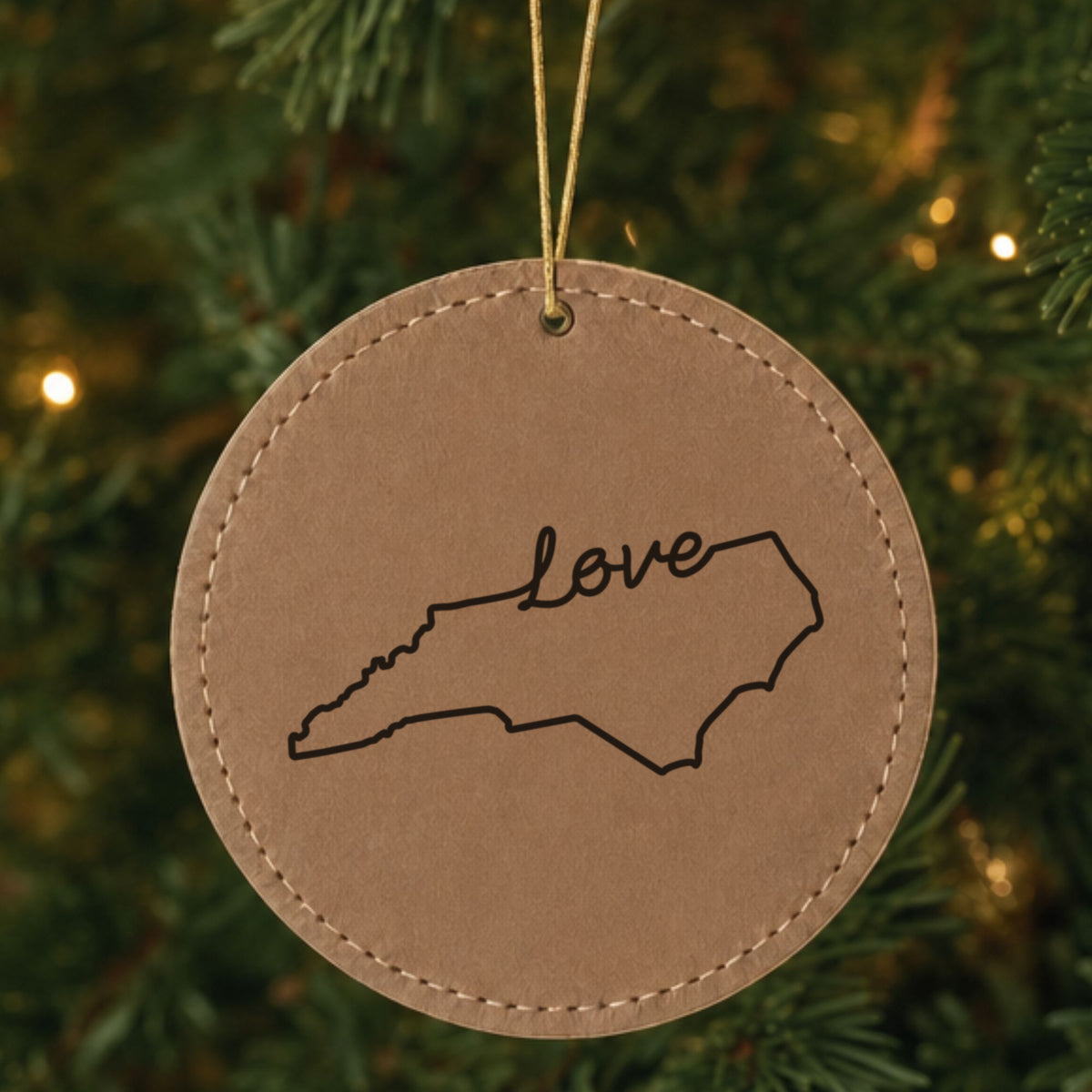 Brown leatherette round Christmas ornament laser engraved with the North Carolina Hand-Drawn Love design, hanging on a tree