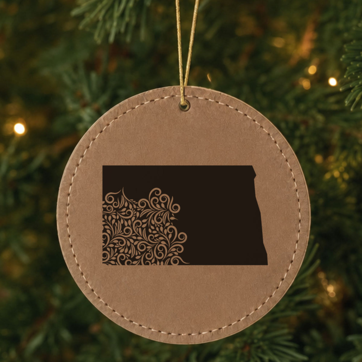 Brown leatherette round Christmas ornament laser engraved with the North Dakota Floral Damask design, hanging on a tree