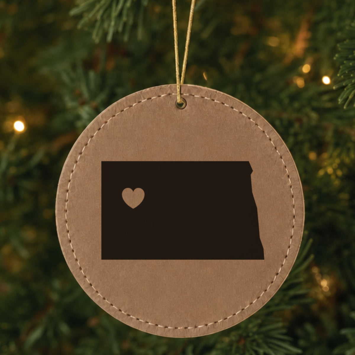 Brown leatherette round Christmas ornament laser engraved with the North Dakota Heart Accent design, hanging on a tree