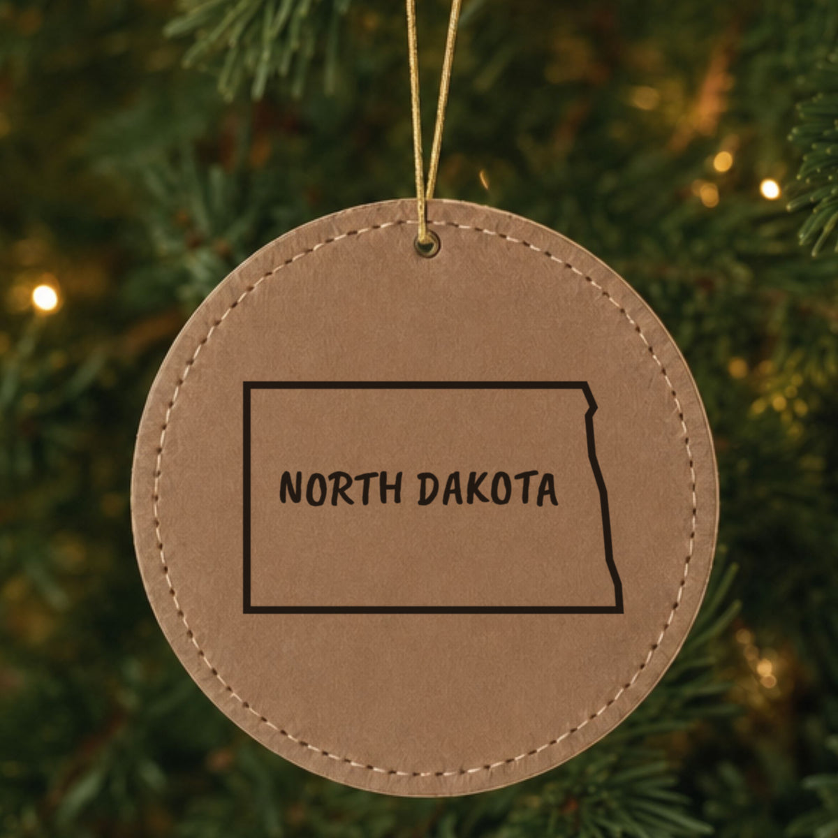 Brown leatherette round Christmas ornament laser engraved with the North Dakota Brush Lettering design, hanging on a tree