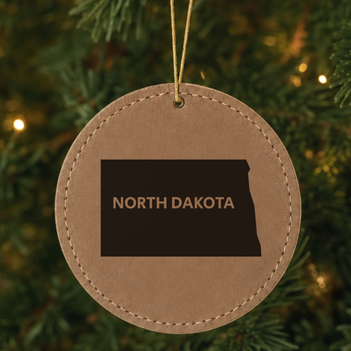 Brown leatherette round Christmas ornament laser engraved with the North Dakota Bold State Name design, hanging on a tree