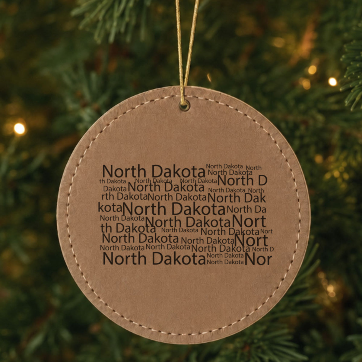 Brown leatherette round Christmas ornament laser engraved with the North Dakota Word Cloud design, hanging on a tree