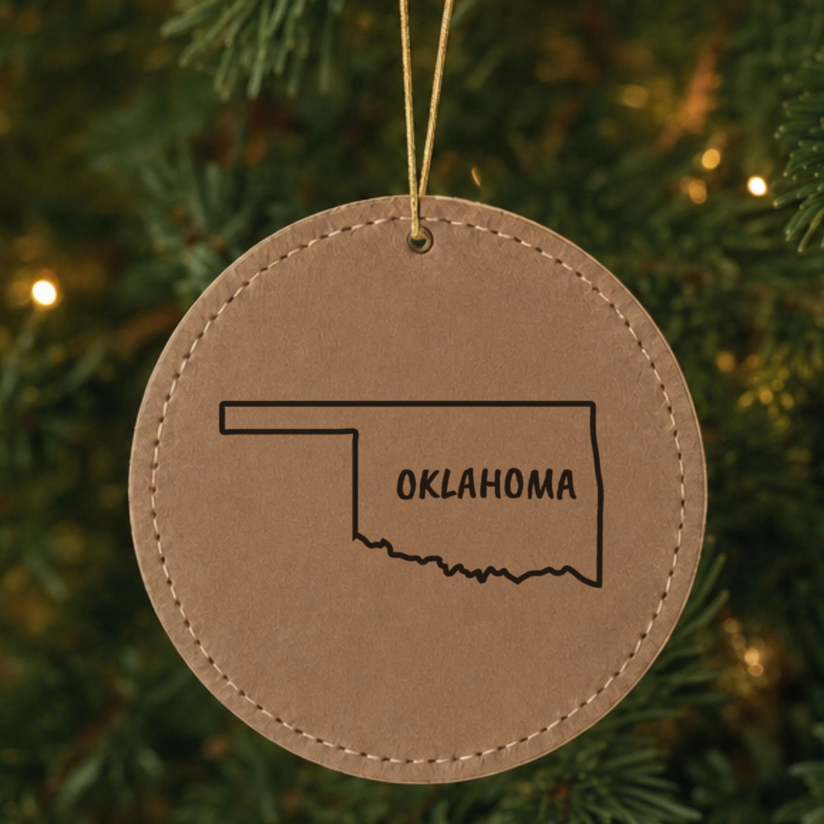 Brown leatherette round Christmas ornament laser engraved with the Oklahoma Brush Lettering design, hanging on a tree