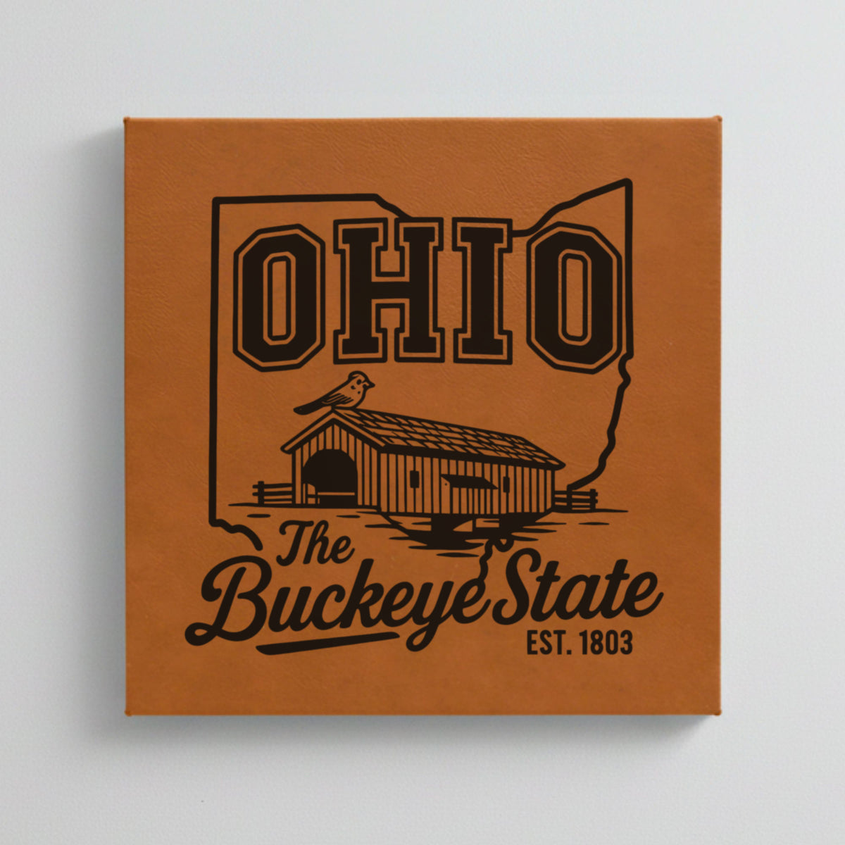 Close up of the Ohio design engraved on Brown leatherette: a covered bridge and a cardinal, "The Buckeye State", EST. 1803