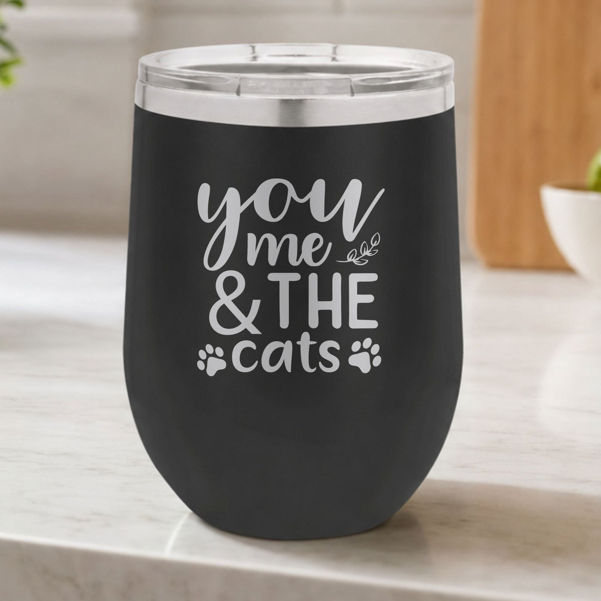 Black 12 oz stemless wine tumbler laser engraved with "You, Me & the Cats", plus paw prints, on a kitchen counter