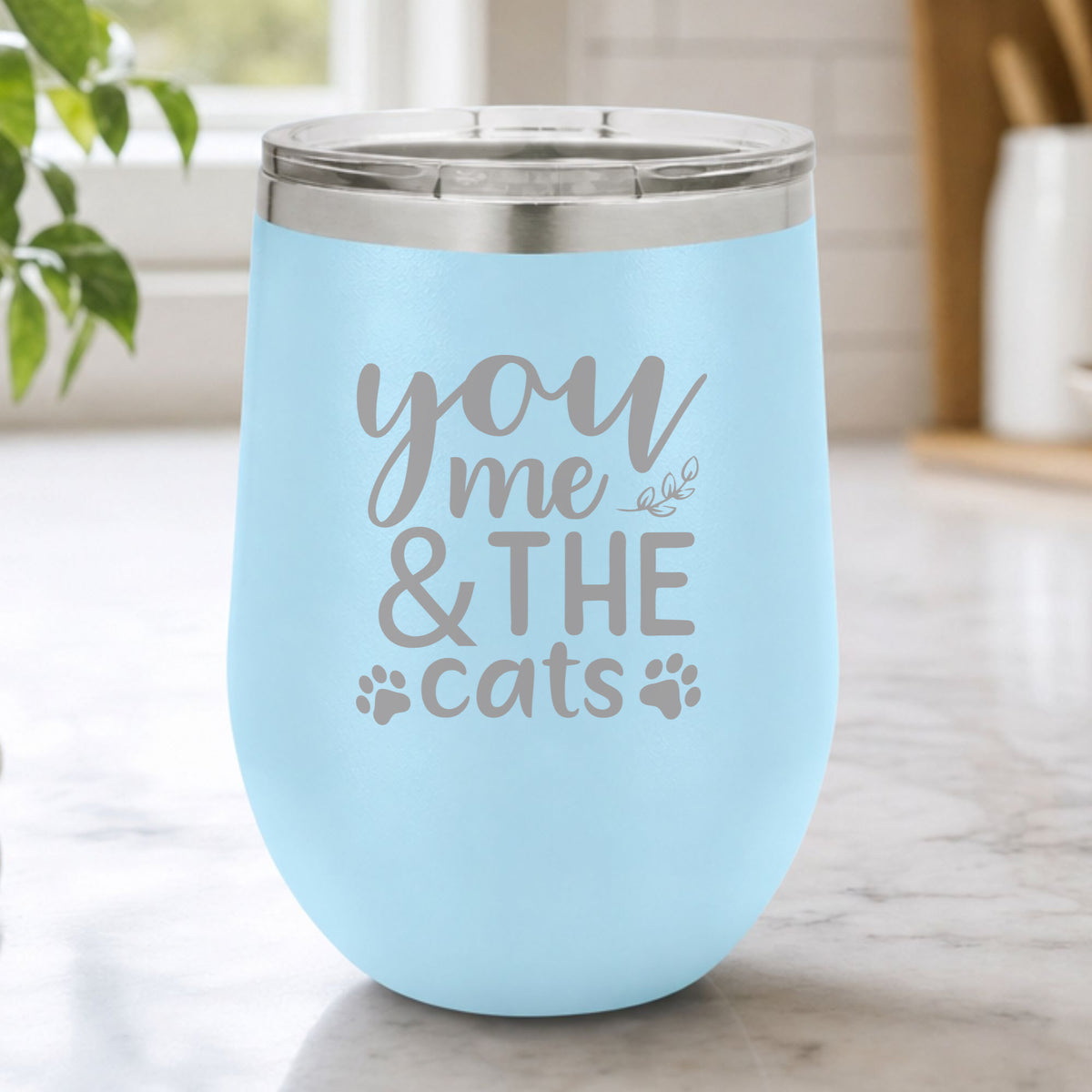 Light Blue 12 oz stemless wine tumbler laser engraved with "You, Me & the Cats", plus paw prints, on a kitchen counter