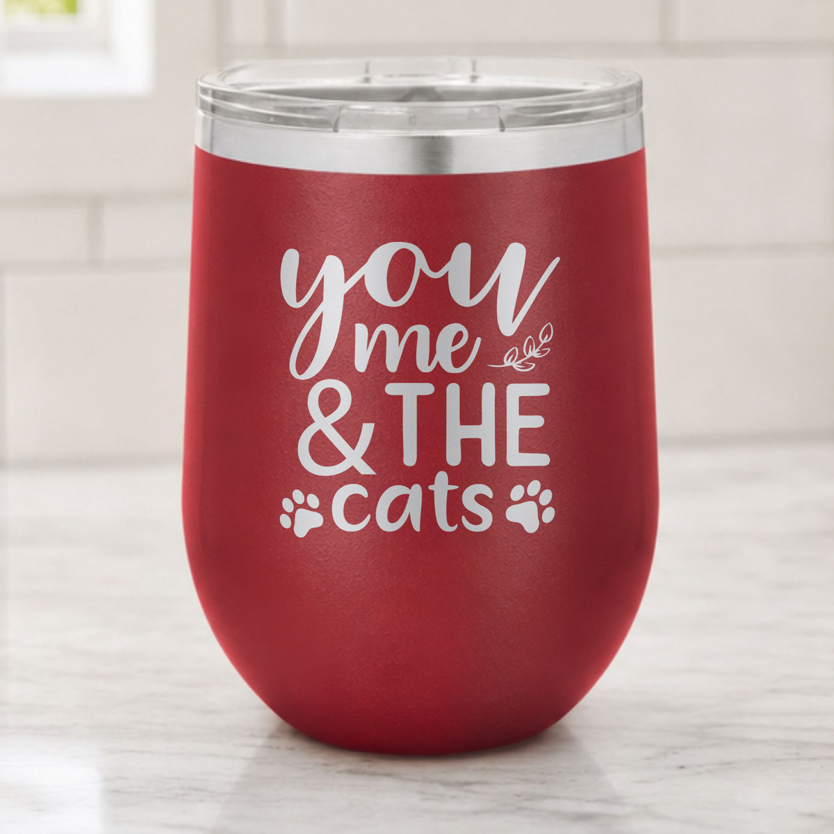 Maroon 12 oz stemless wine tumbler laser engraved with "You, Me & the Cats", plus paw prints, on a kitchen counter