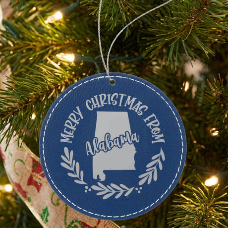 Blue leatherette round Christmas ornament engraved Merry Christmas From Alabama, with the state shape in a laurel wreath, hanging on a tree