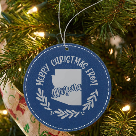 Blue leatherette round Christmas ornament engraved Merry Christmas From Arizona, with the state shape in a laurel wreath, hanging on a tree