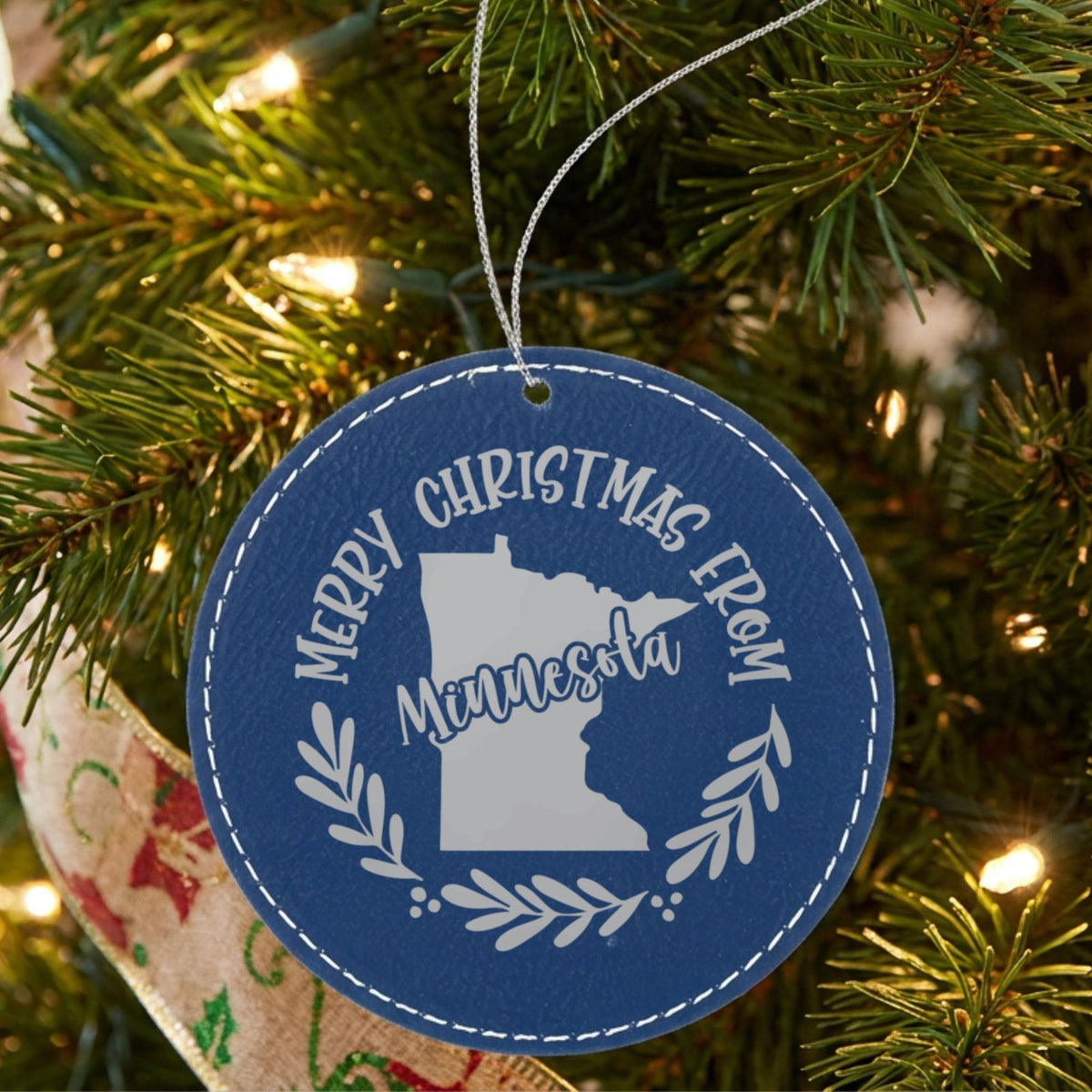 Blue leatherette round Christmas ornament engraved Merry Christmas From Minnesota, with the state shape in a laurel wreath, hanging on a tree