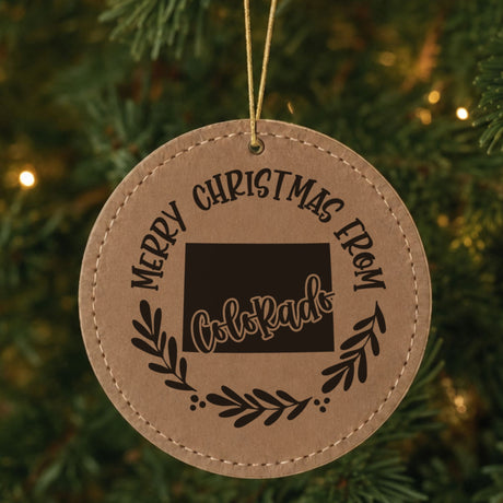 Brown leatherette ornament engraved Merry Christmas From Colorado with the state shape in a laurel wreath, hanging on a tree