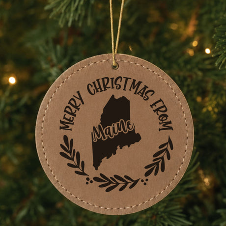 Brown leatherette round Christmas ornament engraved Merry Christmas From Maine, with the state shape in a laurel wreath, hanging on a tree
