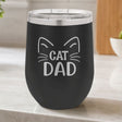 Black 12 oz stemless wine tumbler laser engraved with "Cat Dad" framed by cat ears and whiskers, on a kitchen counter