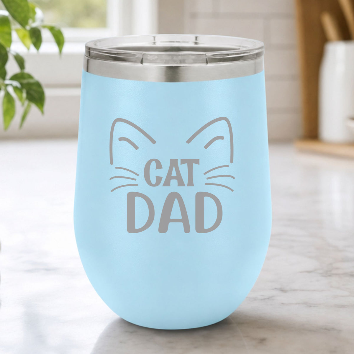 Light Blue 12 oz stemless wine tumbler laser engraved with "Cat Dad" framed by cat ears and whiskers, on a kitchen counter