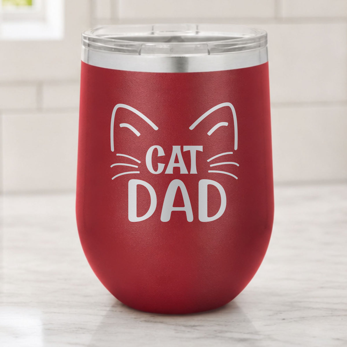 Maroon 12 oz stemless wine tumbler laser engraved with "Cat Dad" framed by cat ears and whiskers, on a kitchen counter