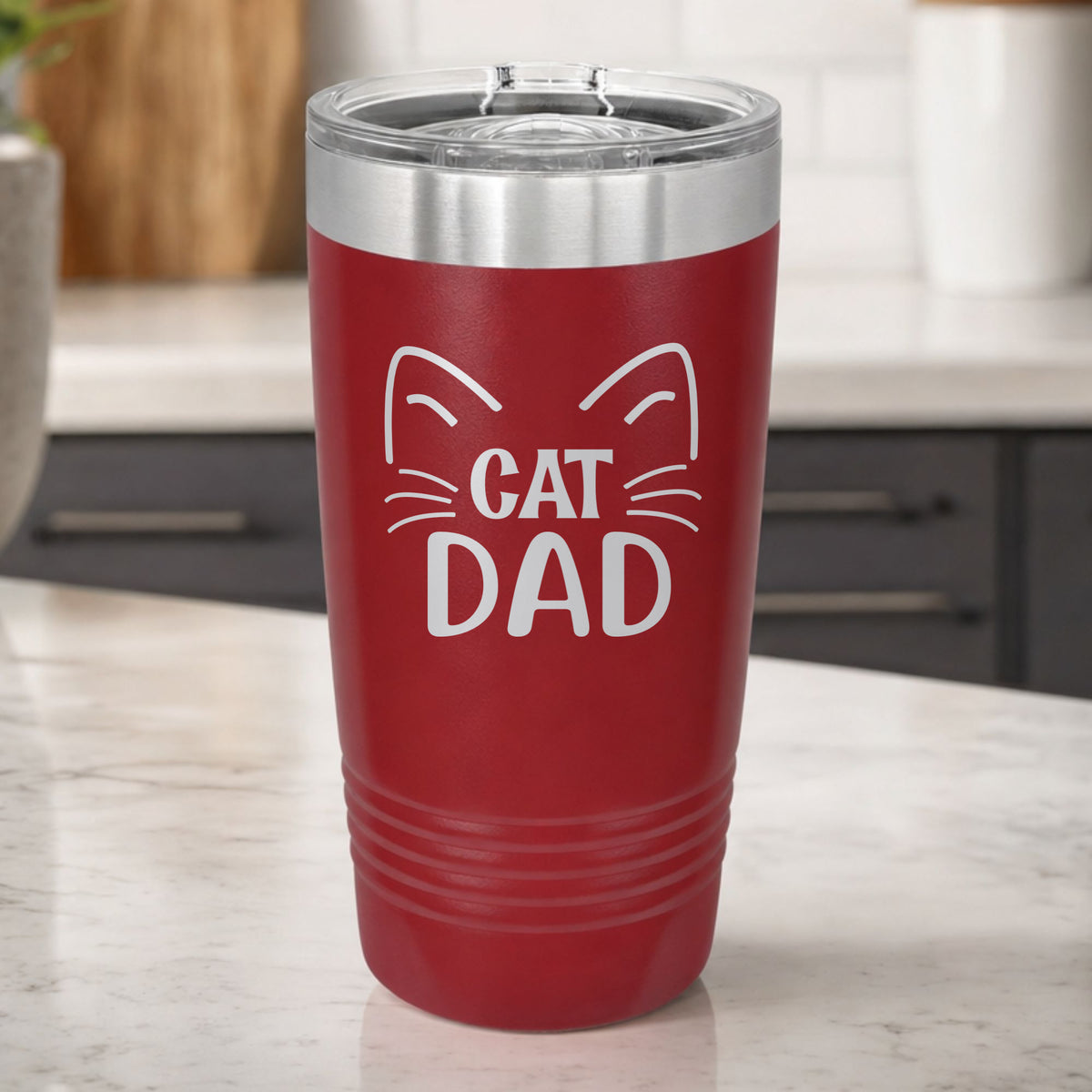 Maroon 20 oz tumbler laser engraved with "Cat Dad" framed by cat ears and whiskers, on a kitchen counter
