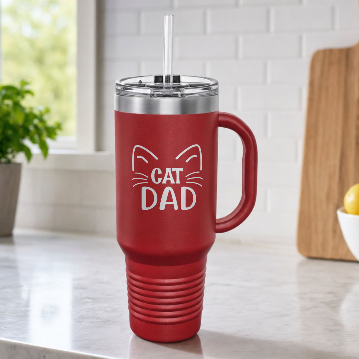 Maroon 40 oz tumbler with handle and straw laser engraved with "Cat Dad" framed by cat ears and whiskers, on a kitchen counter