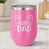 Pink 12 oz stemless wine tumbler laser engraved with "Cat Dad" framed by cat ears and whiskers, on a kitchen counter