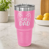 Pink 30 oz tumbler laser engraved with "Cat Dad" framed by cat ears and whiskers, on a kitchen counter