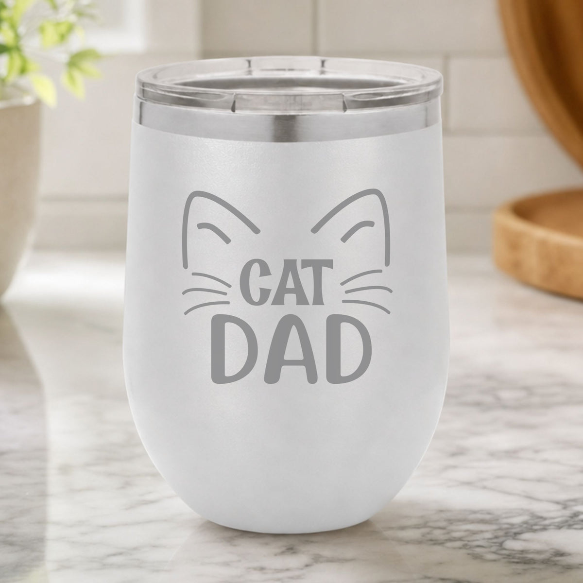 White 12 oz stemless wine tumbler laser engraved with "Cat Dad" framed by cat ears and whiskers, on a kitchen counter