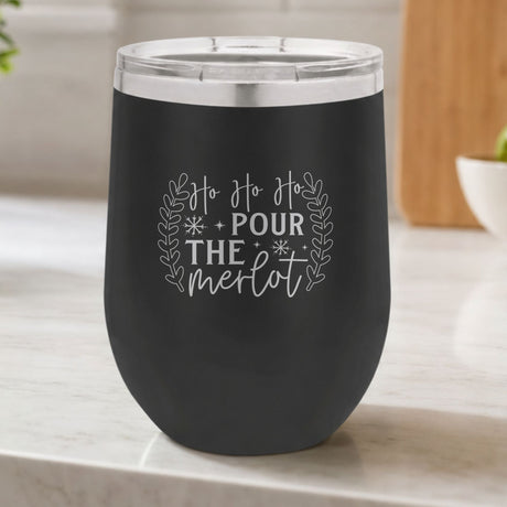 Black 12 oz stemless wine tumbler laser engraved with "Ho Ho Ho, Pour the Merlot", on a kitchen counter