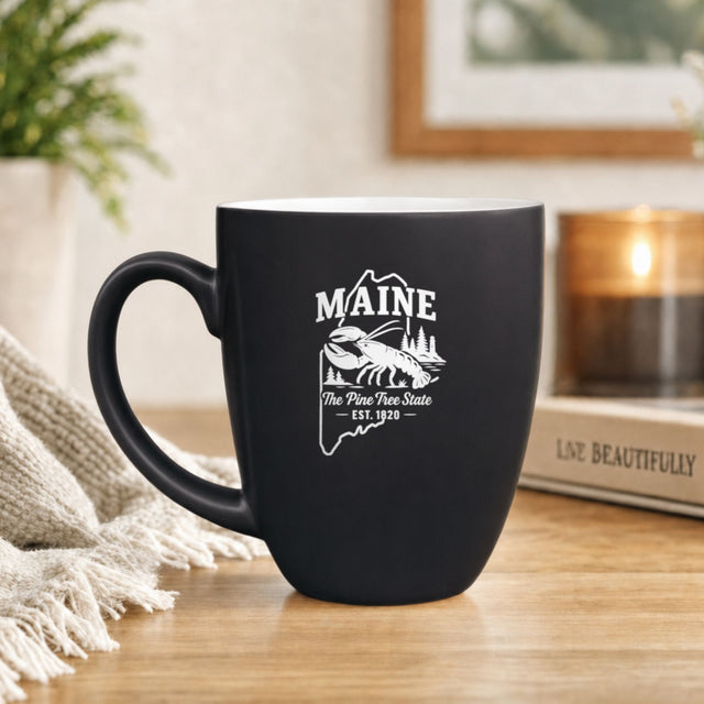 Black 16 oz ceramic coffee mug printed with the Maine state outline and The Pine Tree State