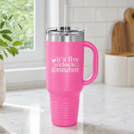 Pink 40 oz tumbler with handle and straw laser engraved with "It's Five O'Clock Somewhere", on a kitchen counter