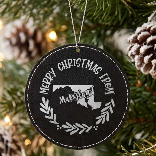 Black leatherette round Christmas ornament engraved Merry Christmas From Maryland, with the state shape in a laurel wreath, hanging on a tree
