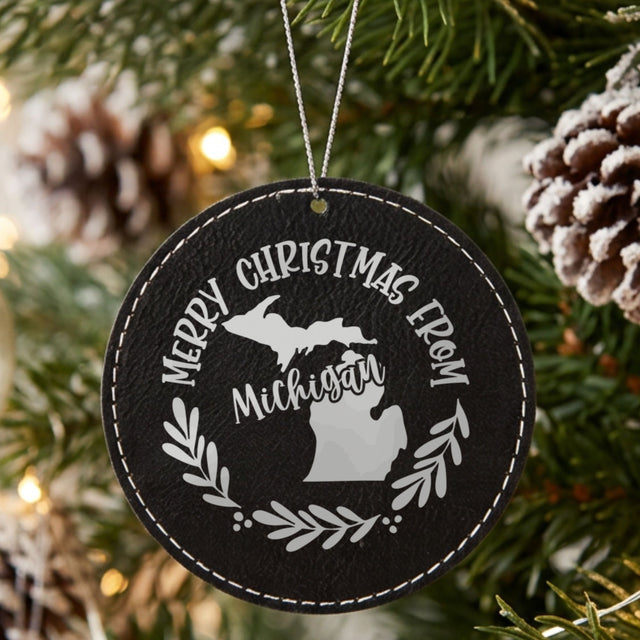 Black leatherette round Christmas ornament engraved Merry Christmas From Michigan, with the state shape in a laurel wreath, hanging on a tree