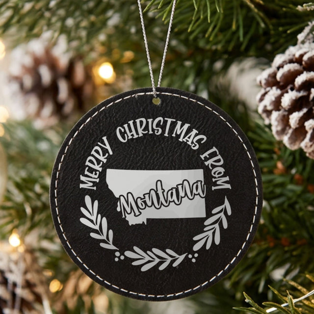 Black leatherette round Christmas ornament engraved Merry Christmas From Montana, with the state shape in a laurel wreath, hanging on a tree