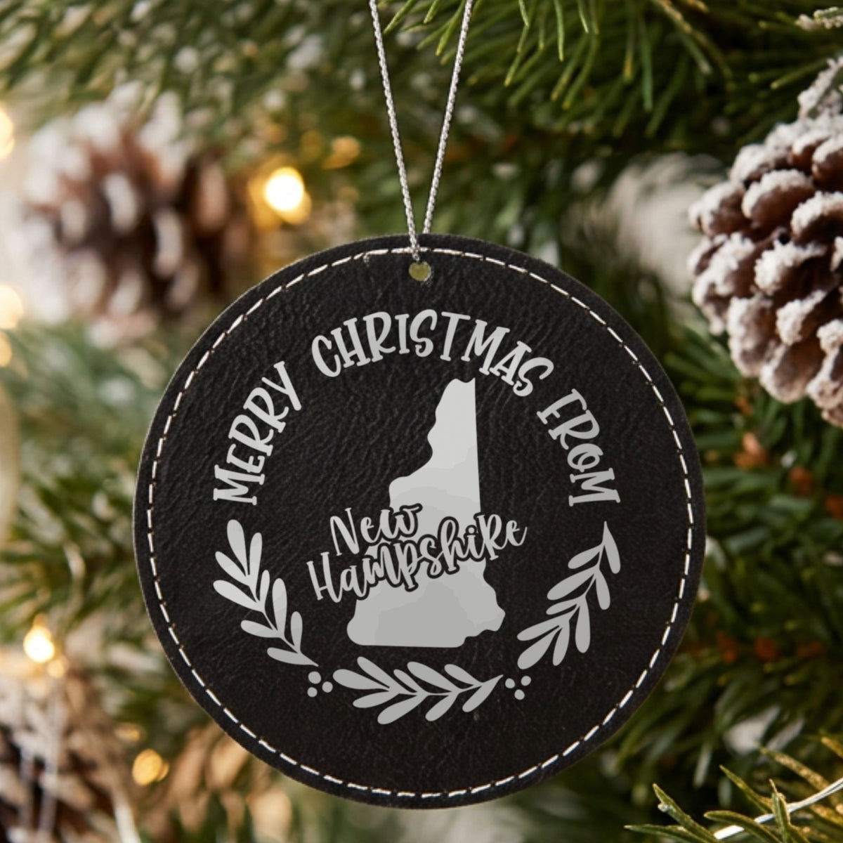 Black leatherette round Christmas ornament engraved Merry Christmas From New Hampshire, with the state shape in a laurel wreath, hanging on a tree