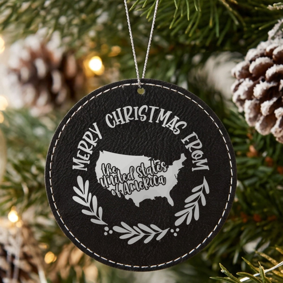 Black leatherette round Christmas ornament engraved Merry Christmas From the USA, with the country shape in a laurel wreath, hanging on a tree